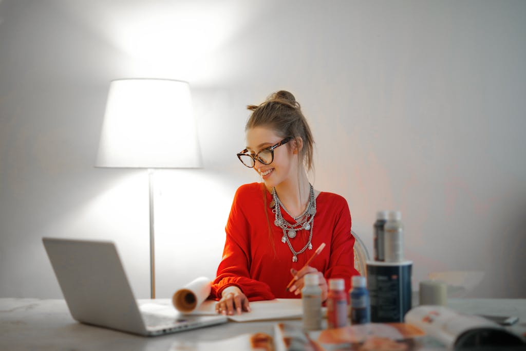 Smiling woman in red using laptop and art supplies at home office desk.