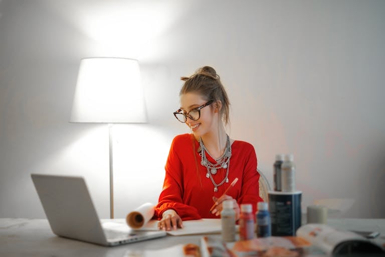 Smiling woman in red using laptop and art supplies at home office desk.